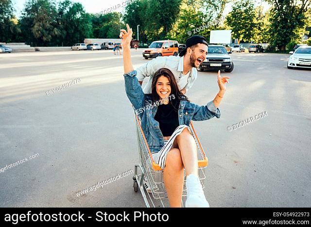 Handsome guy carries his girlfriend in cart in the parking lot at the supermarket