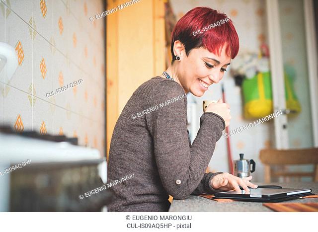 Young woman sitting at table drinking coffee, looking at digital tablet