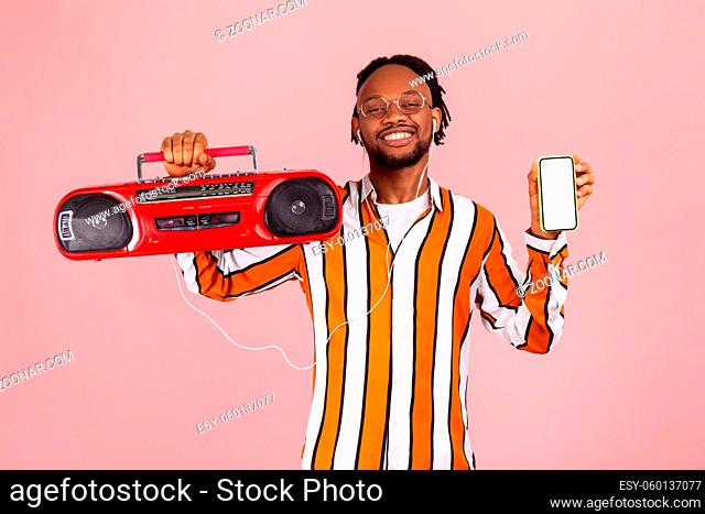 Happy afro-american man with dreadlocks in striped shirt and earphones holding tape recorder and white screen smartphone looking for music app