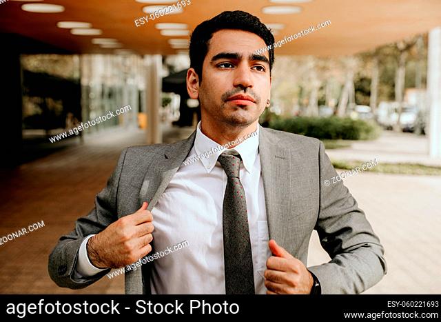 Confident and successful. Handsome young businessman adjusting his jacket and looking away while walking outdoors with office building in the background