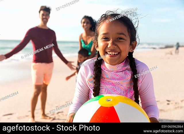 Portrait of smiling biracial girl holding ball with family at beach on sunny day