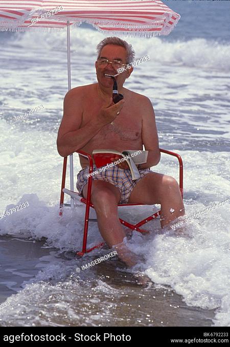 Older man with solar chair and sunshade by the sea, beach, with whistle smoking, holiday, rest, sea waves