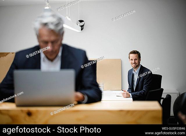 Businessman smiling in office with senior colleague working in foreground