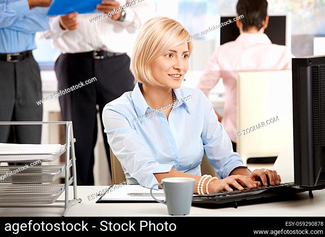 Happy young businesswoman using computer in bright office, colleagues working in the background