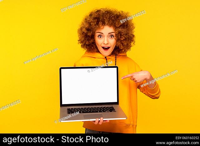 Shocked, surprised woman with fluffy curly hair in urban style hoody pointing at empty laptop screen and looking with amazement, showing mock up