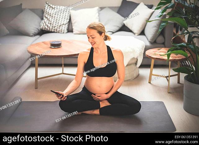 Pregnancy and technoloy supported healthy lifestyle concept. Cheerful happy pregnant woman using smart phone application while exercising on yoga mat on living...