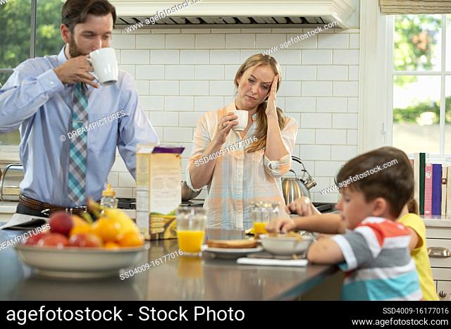 Mom who is feeling under the weather with a headache with family in kitchen, struggling to get the kids breakfast and ready to start the day