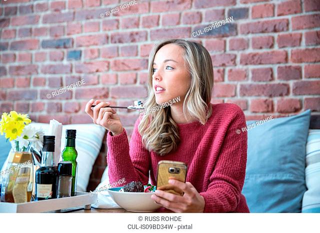Woman sitting in cafe, eating bowl of muesli, holding smartphone
