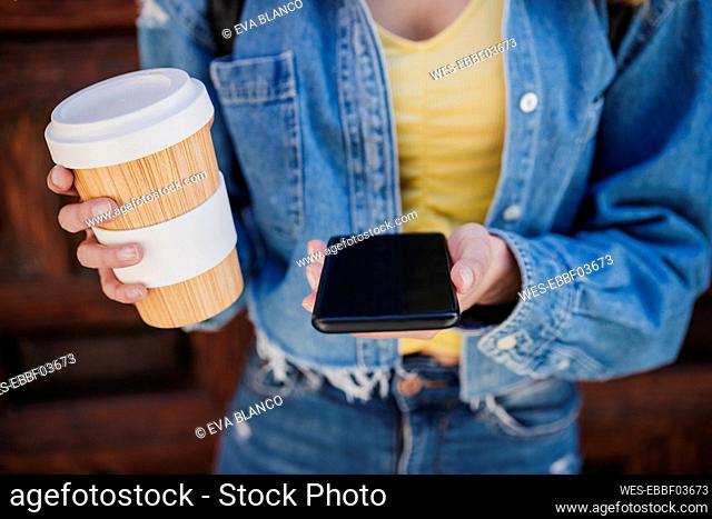 Woman holding reusable cup and smart phone outdoors