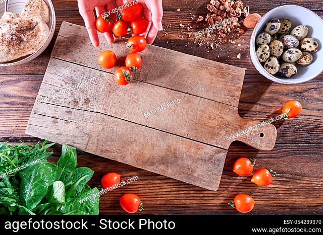 A set of ingredients quail eggs, fresh herbs, nuts, boiled meat on a wooden table. The hands of the girl put tomatoes on the kitchen board with a copy space