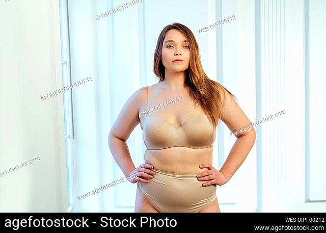 Confident curvy model in beige lingerie with hand on hip standing against white wall