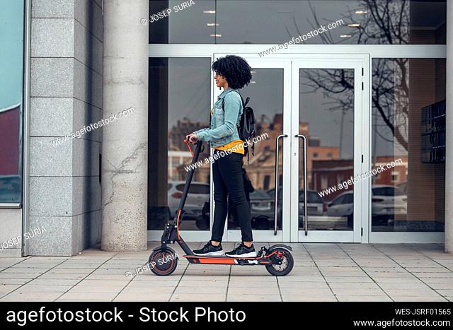 Woman riding electric push scooter on footpath near building
