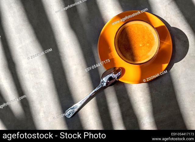Latte drink in the glass on the orange saucer on the table. There is a chrome spoon near it. Sun shines onto them and creates glares and shadow patterns