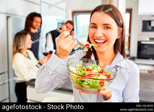 Junge Frau beim Salat essen in WG Küche vor einer Gruppe Freunde