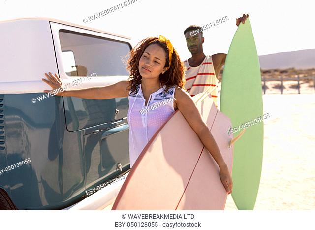 Front view of young Mixed-race couple with surfboard leaning on camper van at beach