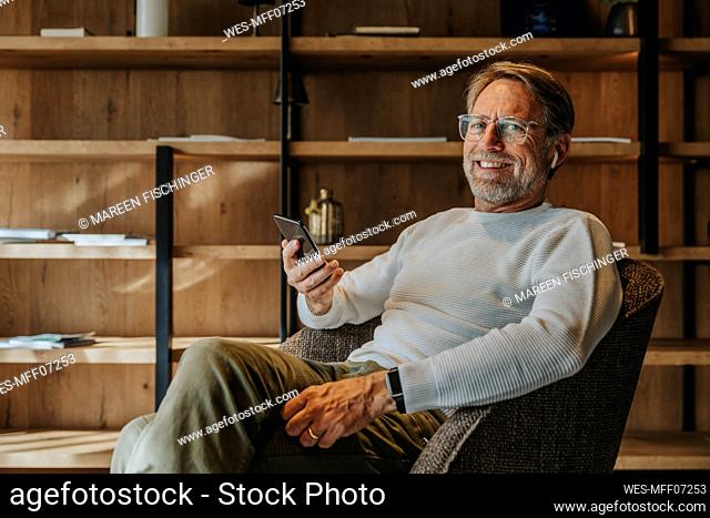 Smiling mature man holding mobile phone while sitting on armchair