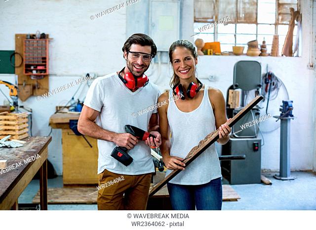 Male and female carpenters standing together with wooden plank and drill machine