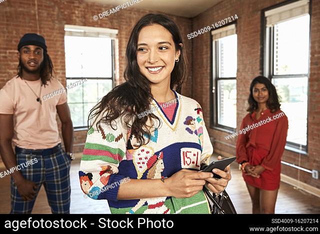 Group of friends in empty loft space, socially distanced group photo, focus on woman in foreground holding tablet looking off camera