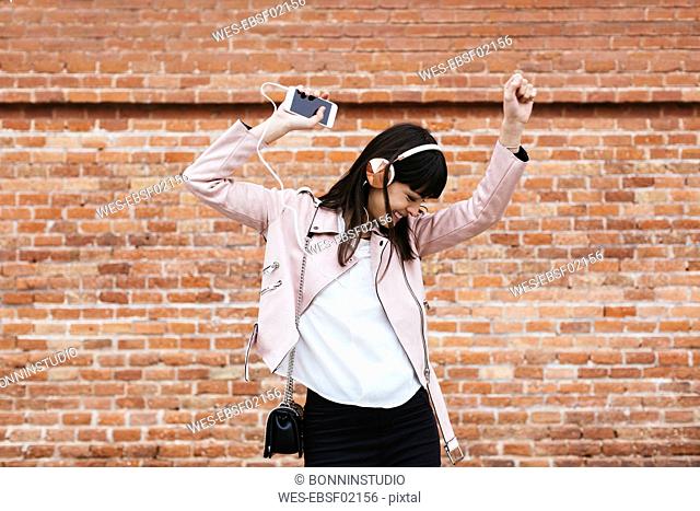 Happy woman with cell phone listening to music on headphones at brick wall