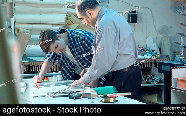 Senior male worker instructing young trainee how to cut a glass for frame behind the desk in frame workshop. Professional shot in 4K resolution. 083