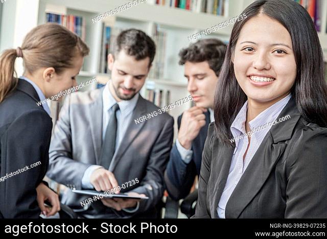 Business woman smiling and looking at the camera with her colleagues talking and looking down at a digital tablet in the background