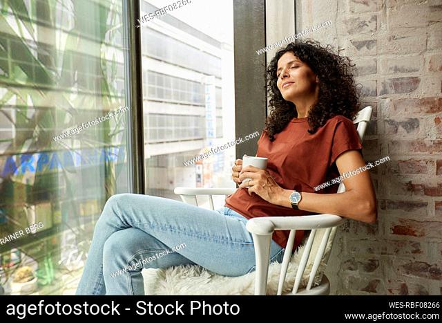 Relaxed businesswoman holding coffee cup while sitting with eyes closed on chair by window