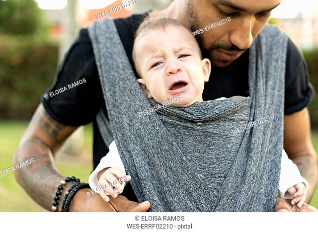 Young father carrying his crying baby son in a baby sling