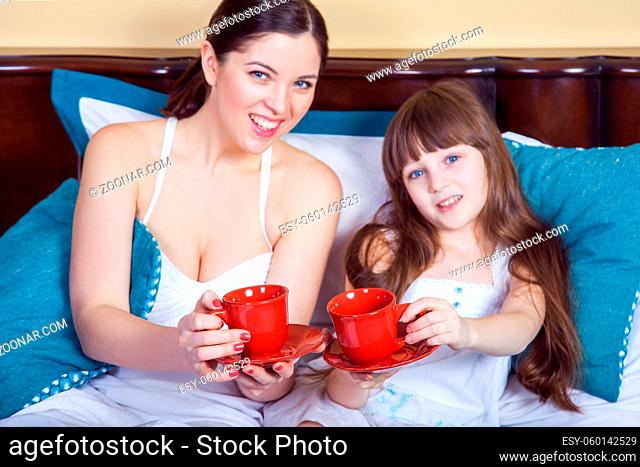 Happy mother and daughter having fun and holding cup and resting in bed, looking at camera with toothy smile