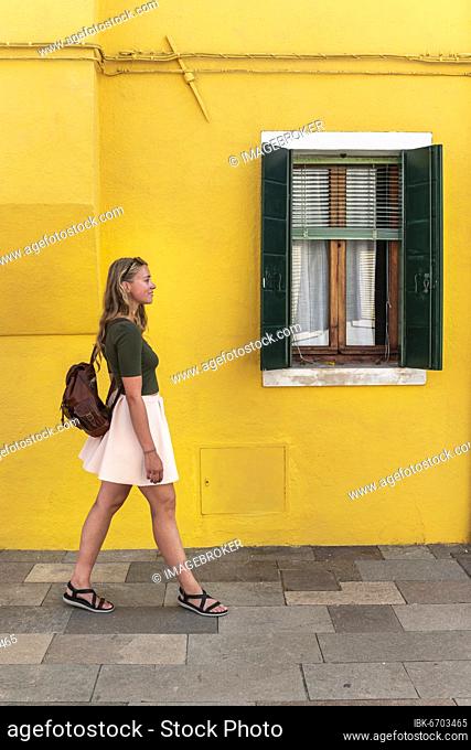 Young woman walking in front of colorful house, yellow house facade, tourist on Burano island, Venice, Veneto, Italy, Europe