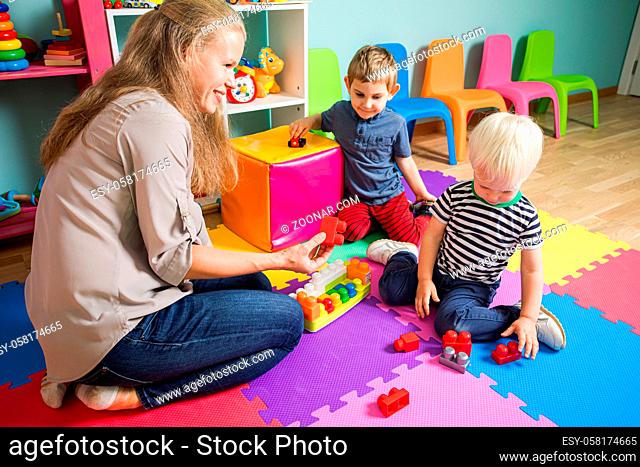 Two little boys are playing with a kindergarten teacher. The woman is sitting with boys on the floor in the playroom