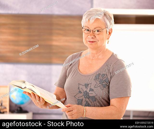 Portrait of senior teacher standing in classroom, holding book, looking at camera