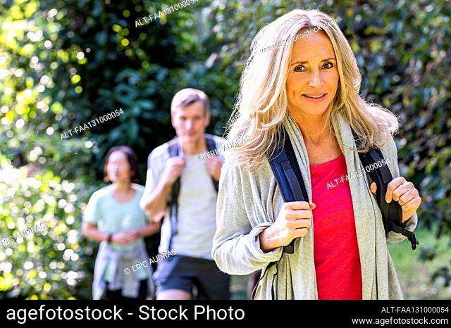 Mid-shot of blonde middle-age woman with backback hiking with her diverse group of friends in the woods
