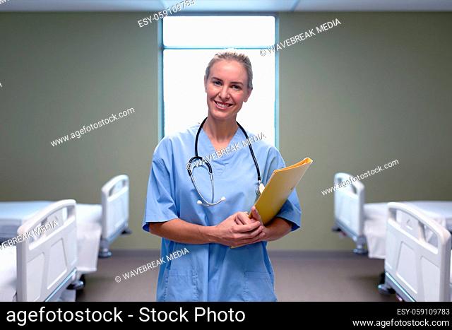 Portrait of smiling caucasian female doctor in hospital wearing scrubs and stethoscope holding files