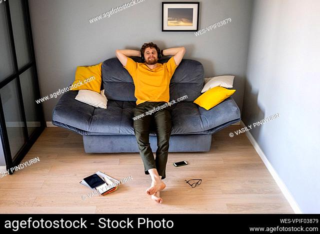Man wearing headphones relaxing with hands behind head on sofa at home