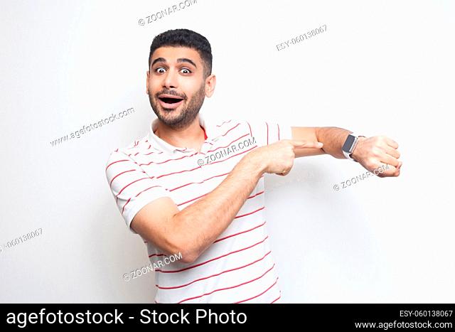 Time to go. Portrait of amazed funny bearded young man in striped t-shirt standing, pointing at his smart watch, looking at camera with surprised face