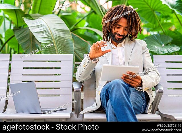 Smiling businessman using tablet PC sitting by laptop on chair