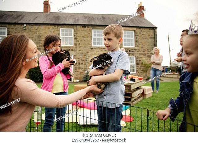 Young boy standing outdoors, a mid adult woman is handing him a bubby rabbit to hold