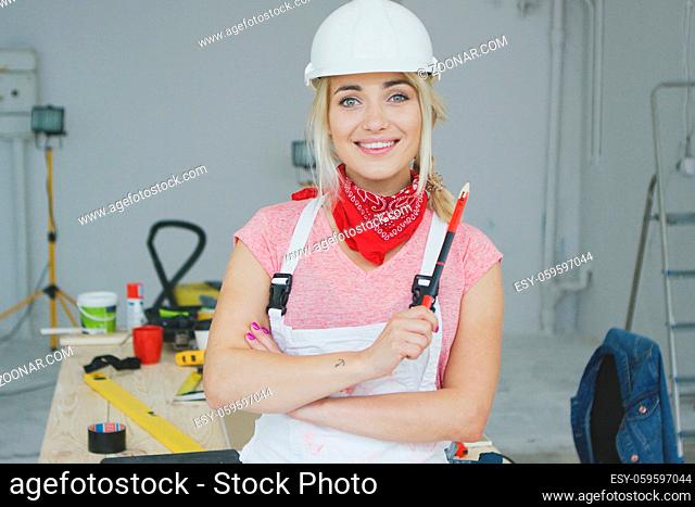 Beautiful young blond woman in white safety helmet and overalls with red bandana around neck looking at camera and smiling standing with pencil at workplace...