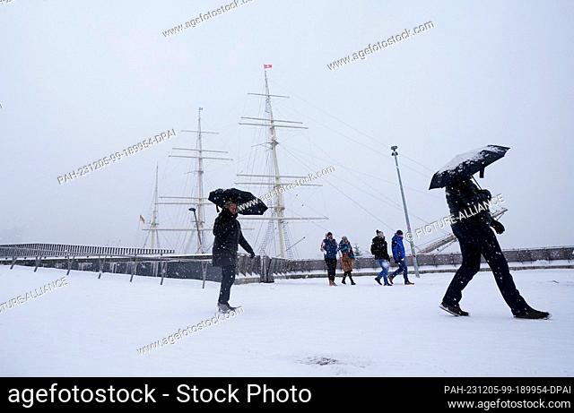 05 December 2023, Hamburg: People with umbrellas walk past the Rickmer Rickmers museum ship in the harbor in a snowstorm