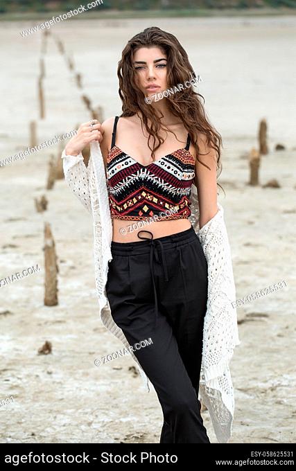 Beautiful girl with crossed legs stands on the sand outdoors. She wears black pants, multi-colored top with patterns and a white cardigan