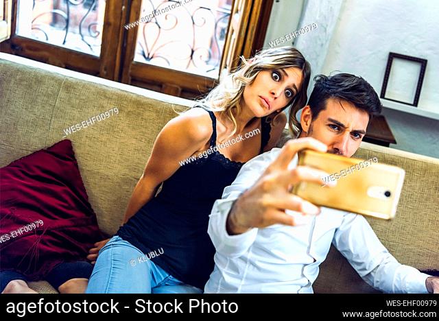 Young couple making faces while taking selfie on couch in living room at home