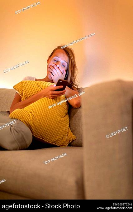 Cute young woman on a comfortable sofa in her modern apartment using her cell phone - staring at the blue lkight emitting device instaed of getting a good night...