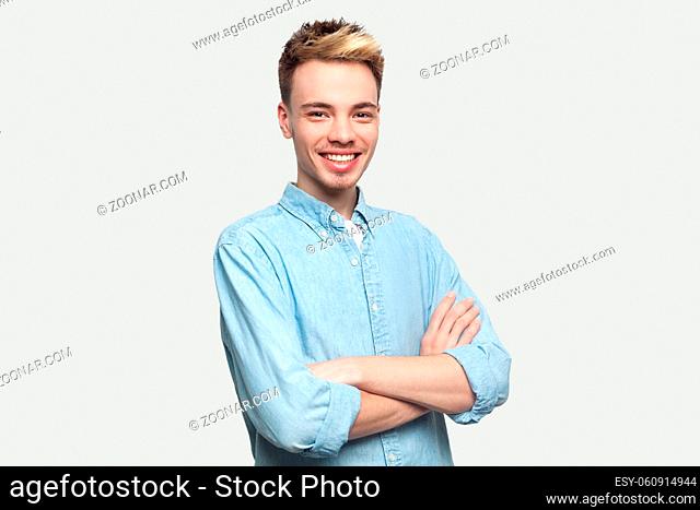 Portrait of happy satisfied handsome young man in light blue shirt standing with crossed arms and looking at camera with toothy smile