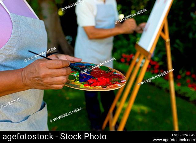 Midsection of biracial senior wife holding palette and paintbrush while man painting in background