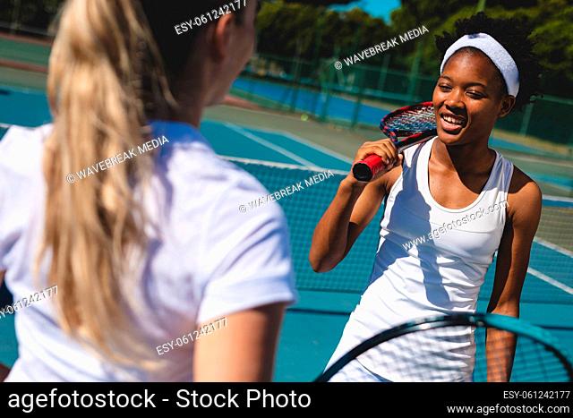 Smiling african american female player talking with caucasian athlete at tennis court on sunny day