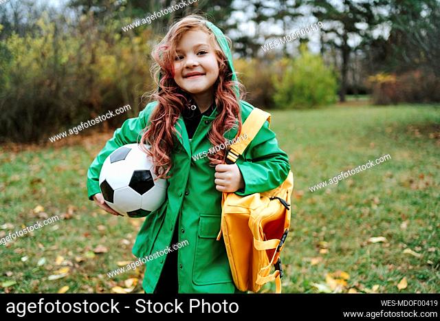 Smiling girl standing with backpack and soccer ball at park
