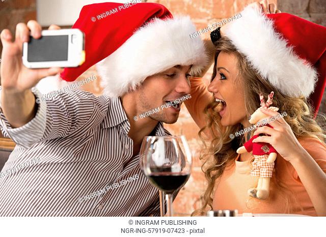 Young happy couple wearing Santa hats looking at one another in restaurant, kissing and taking photos of themselves on mobile phone camera while celebrating...