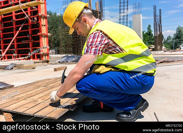 Side view of a young worker wearing safety vest and yellow hard hat while hammering a nail into wood, during work on the construction site of a contemporary...