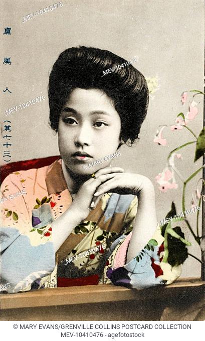 A Pretty young Japanese girl in a floral kimono, posing seated at a table alongside some orchids