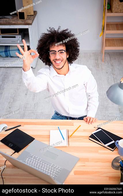Freelance or business concept. Hipster freelance man in glasses showing yo or okay sign to camera while sitting at table and working on laptop computer at home